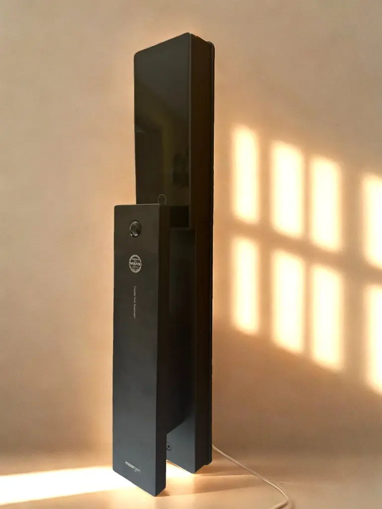 La imagen muestra una cerradura inteligente de diseño minimalista y moderno, compuesta por dos paneles verticales de acabado negro mate o gris oscuro, con líneas rectas y estilizadas. En el panel principal destaca el logo de la marca "moorgen" en la parte inferior y un pequeño sensor o lector en la sección superior, posiblemente para huellas o reconocimiento. También incluye un texto vertical relacionado con su funcionalidad, lo que resalta su sofisticación tecnológica. Está iluminada por una cálida luz natural que proyecta sombras en el fondo beige, destacando su estética premium y funcionalidad para espacios contemporáneos.