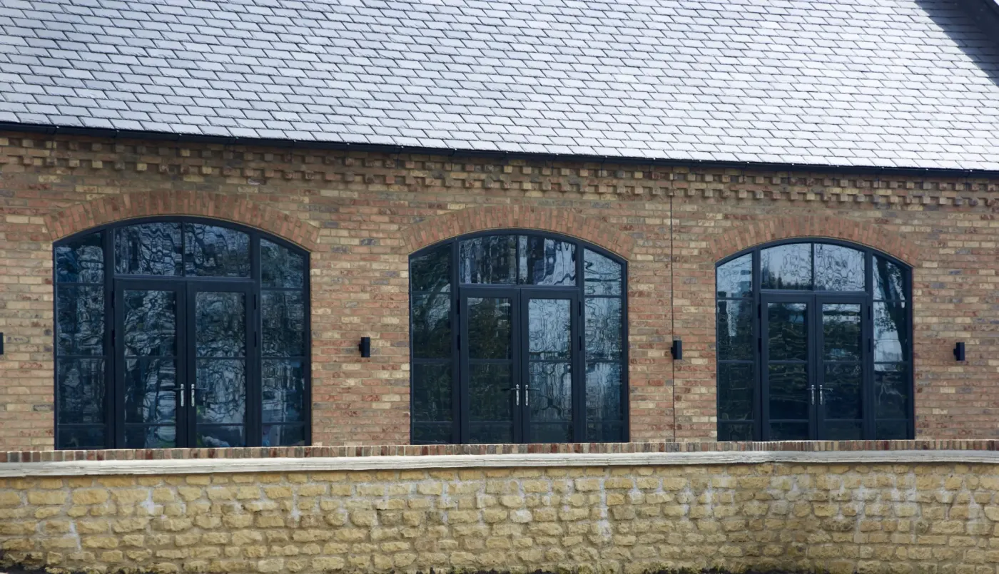 Ventanas y puertas de estilo crittall con arco y retícula negra en la fachada de piedra y ladrillo de una casa en Girona